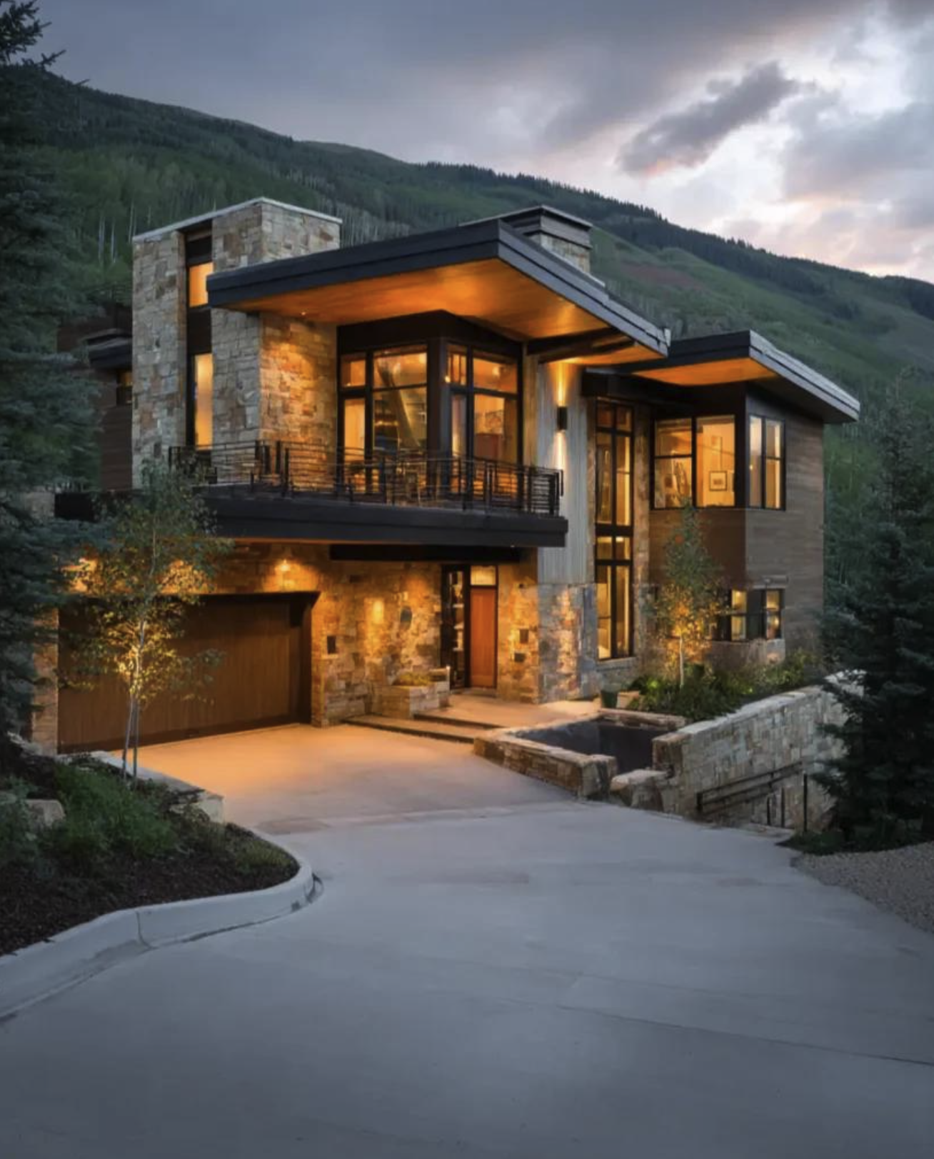 A contemporary stone-and-timber mountain residence glowing at dusk below a forested ridge